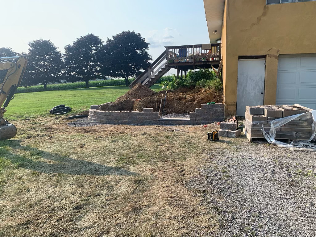 Excavator setting the first courses of a block retaining wall below a deck