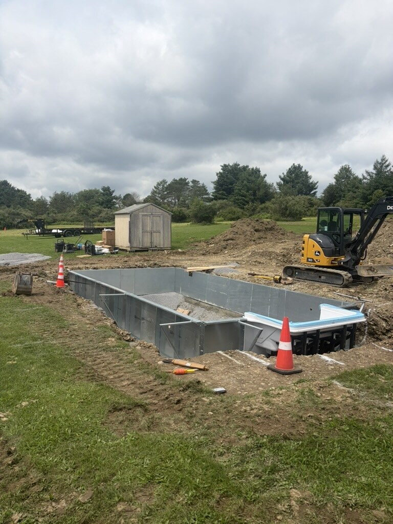 Braced steel pool walls in a freshly excavated yard with a John Deere mini excavator beside them