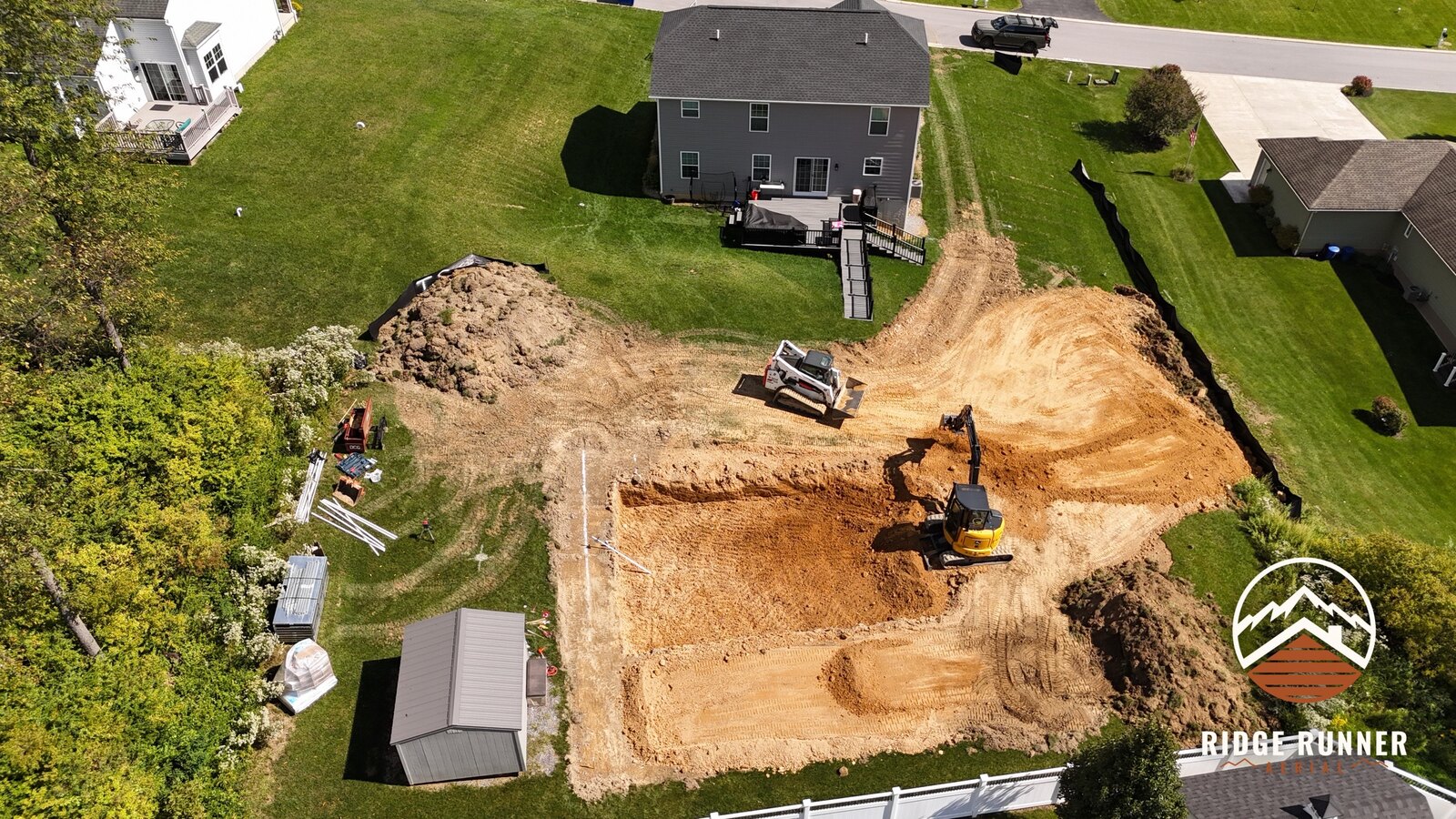 Drone shot of the pool dig from above the deck, excavator swinging spoil to the stockpile in Hollidaysburg, PA